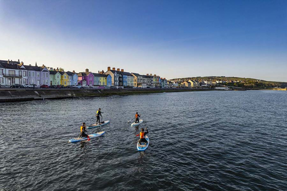 A group of stand-up paddle-boarders in the sea off the coast of Whitehead with its colourful houses in the background